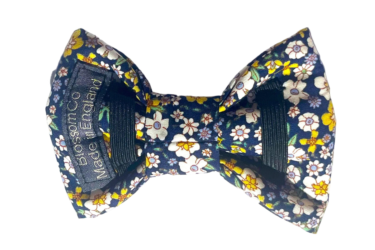 The Agnes  pre-tied dog bowtie features a navy blue fabric with a white, yellow, and green floral print, secured onto the dog collar with two black elastic bands.