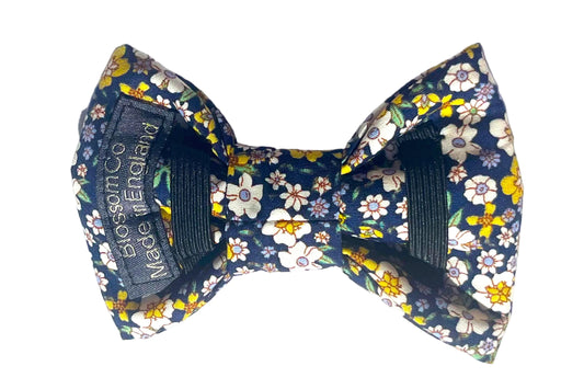 The Agnes  pre-tied dog bowtie features a navy blue fabric with a white, yellow, and green floral print, secured onto the dog collar with two black elastic bands.