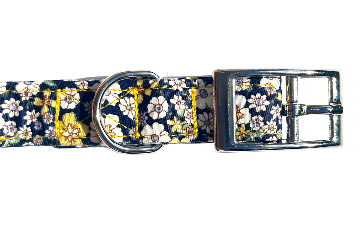 A close-up of BlossomCo's Agnes dog collar with a navy blue background, featuring a white and yellow floral pattern. Image highlights the metal D-ring and buckle.